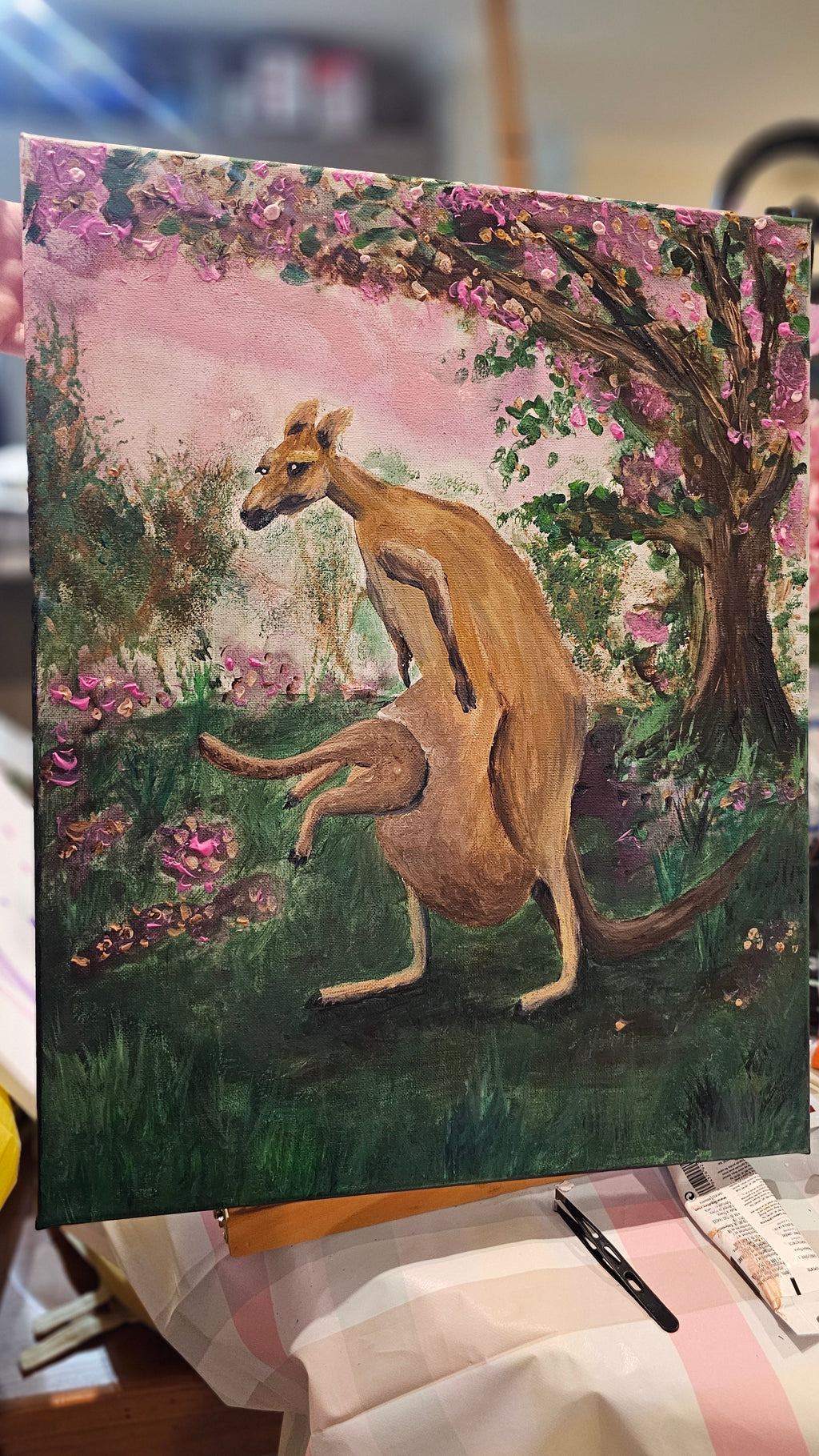 Original Painting by Shelley Bailey of a kangaroo in a garden with flowers and trees. Joey bounds into mother's pouch.